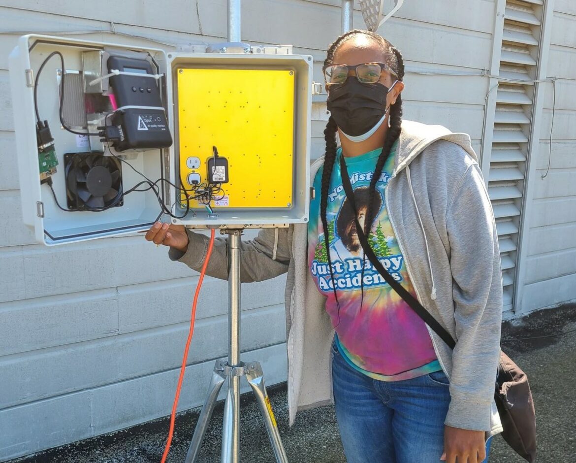 Volunteer Tiffany Williams displays an open box filled with electrical air monitoring equipment.