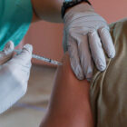 A gloved provider holds a needle to a person's shoulder in preparation for a vaccine injection.