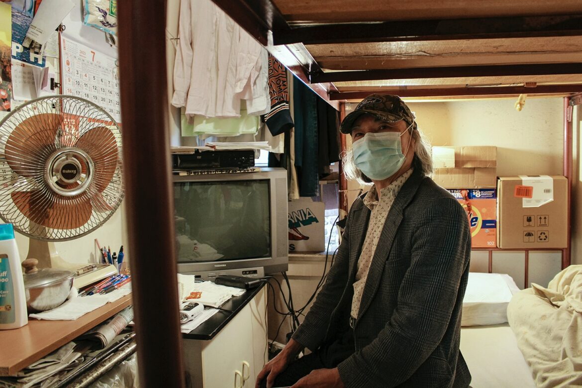 A man in his eighties sits on his bed in a small single-room occupancy residence in Chinatown. Behind him is a window where he hangs his clothes. There are assorted items beside him, including a desk fan, a television, newspapers and cardboard boxes.