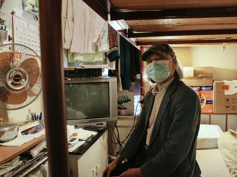 A man in his eighties sits on his bed in a small single-room occupancy residence in Chinatown. Behind him is a window where he hangs his clothes. There are assorted items beside him, including a desk fan, a television, newspapers and cardboard boxes.