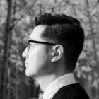 A black-and-white profile photo of a man facing left in front of leafy trees.