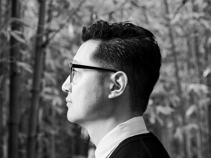 A black-and-white profile photo of a man facing left in front of leafy trees.