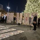 Several people stand in front of an evergreen tree covered in small white lights. They are holding vertical white banners displaying lists of people's names in colorful letters. Other banners are spread on the ground in front of them. A woman with shoulder-length gray hair wearing a black coat addresses the crowd speaking into a microphone on a stand.