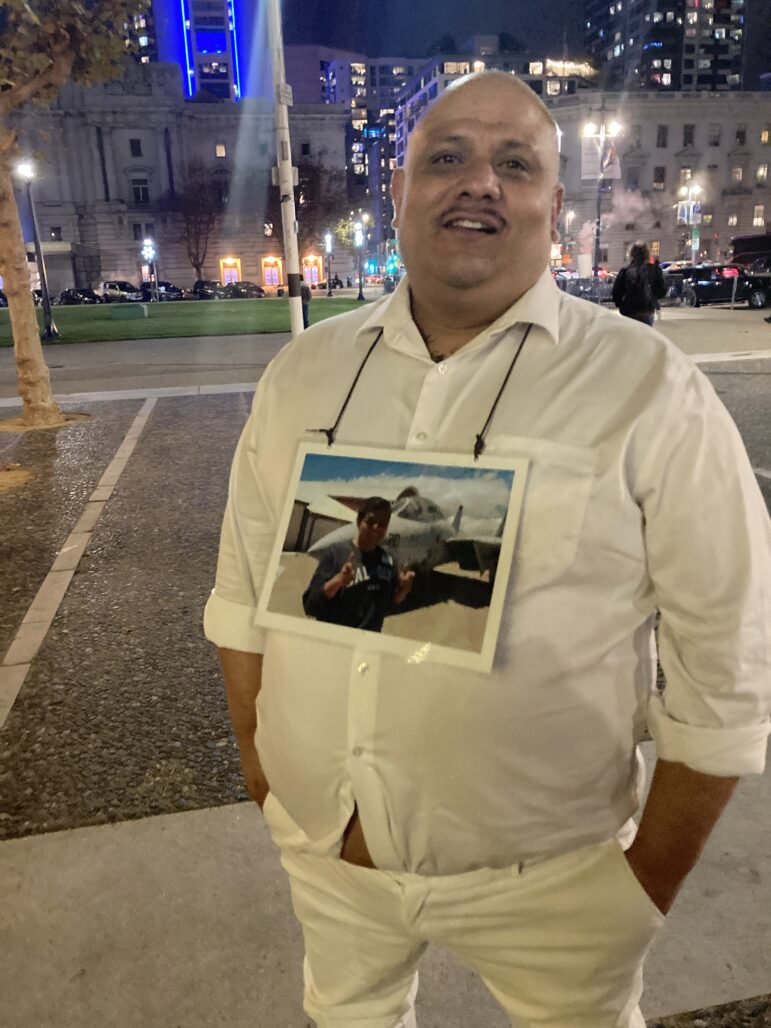 A middle-aged Latino man faces the camera wearing white pants and a white button-down shirt. His hands are in his pockets and he wears on a cord around his neck a large photograph of a young man posing in front of a military jet.