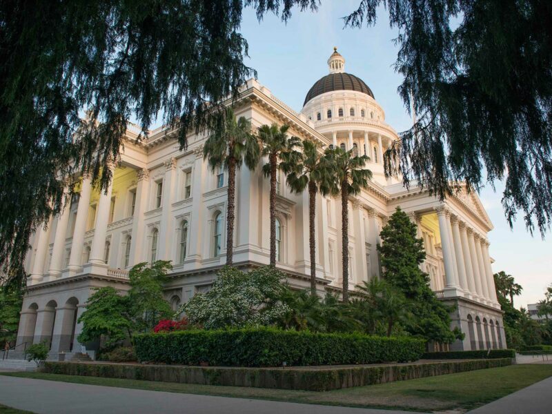 Exterior image of the California State Capitol building in Sacramento. Taken from a corner of the building at a low angle, the cream colored facade features columned entrances on each side, with the building's stately dome appearing toward the top of the frame. The building is surrounded by lush green landscaping.