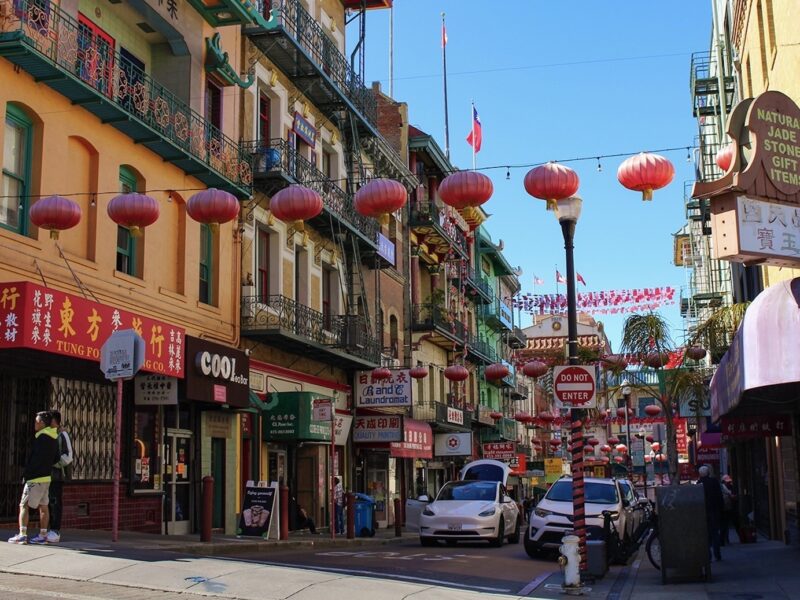 Red lanterns and flags are strung across the roadway on a block in San Francisco's Chinatown. Most of the three and four-story buildings have shops on the ground floor and apartments or offices above. Many of them have wrought iron balconies that are painted green.