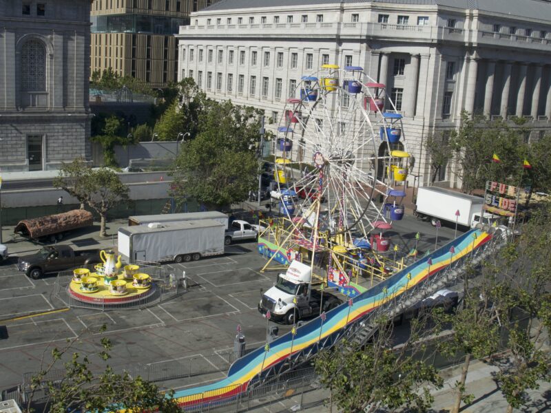 Three colorful carnival attractions — a Ferris wheel, a wavy slide and a tea cup ride — are being set up on a street in San Francisco's Civic Center neighborhood. Several trucks are parked nearby.