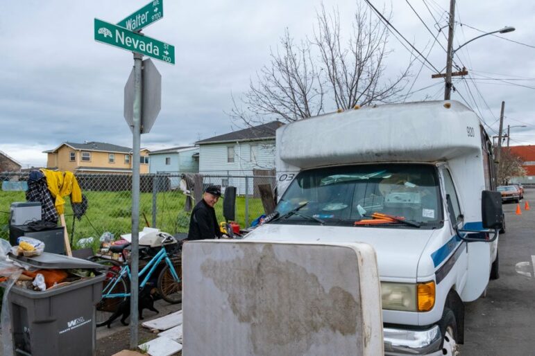 A mattress leans against the front of a small white bus with a trashcan, bicycle, clothing and other personal items on the sidewalk and hanging on a nearby fence.