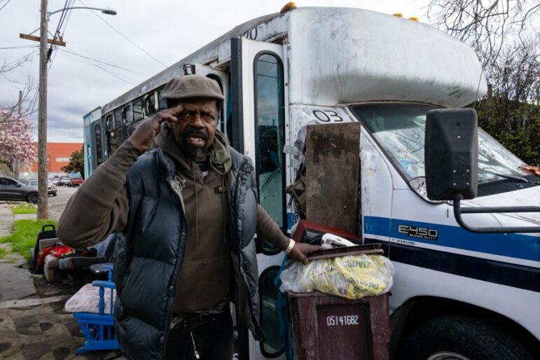 A man wearing a cap and a puffy navy vest holds one hand near his forehead and the other on the lid of a trashcan as he stands near a small white bus.