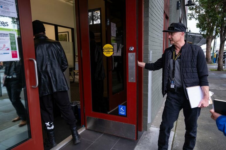 A man holding a clipboard and wearing a navy cap holds open a glass door with red trim to allow someone to enter the building ahead of him.