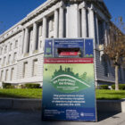 A metal ballot box covered with colorful decals featuring election information is located on a sidewalk in front of a green lawn with the tall columns of San Francisco's City Hall in the background.