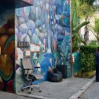 A man in a white shirt holding a skateboard speaks to another man on the sidewalk. In front of a colorful building nearby, a grey office chair rests next to a black trash bag and suitcase.