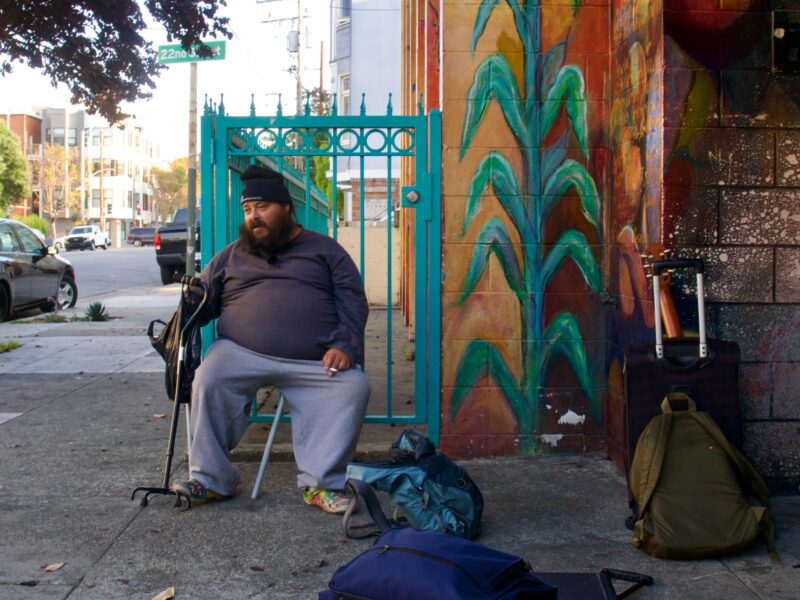 A man in gray sweatpants, a long sleeved shirt and a black beanie sits on a chair with his hand on his cane. To his left, several backpacks and suitcases are scattered on the sidewalk.