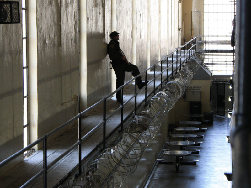 A prison guard stands inside a hall at San Quentin State Prison, his leg up on a railing fortified with razor wire, overlooking the ground floor.