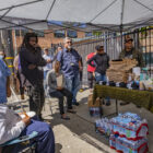 People stand and sit under and around a white shade structure. There is a table stacked with pizza boxes and other snacks, with three large bundles of bottled water underneath.