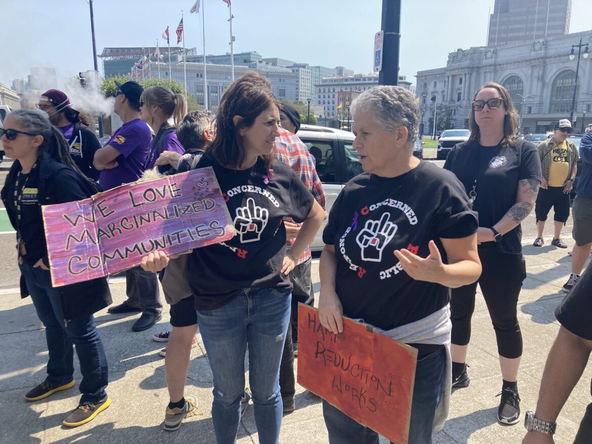 Two women are speaking to each other at a protest rally on a plaza outside of San Francisco City Hall. They are Supervisor Hillery Ronen, who holds a pink sign that reads "We Love Marginalized Communities," and Vitka Eisen, CEO of HealthRight 360, who holds a sign that reads "Harm Reduction Works." They are both wearing jeans and black T-shirts. Many of the people standing around them are similarly dressed.