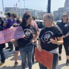 Two women are speaking to each other at a protest rally on a plaza outside of San Francisco City Hall. They are Supervisor Hillery Ronen, who holds a pink sign that reads "We Love Marginalized Communities," and Vitka Eisen, CEO of HealthRight 360, who holds a sign that reads "Harm Reduction Works." They are both wearing jeans and black T-shirts. Many of the people standing around them are similarly dressed.