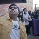 Still from "Homeroom" in which student Denilson Garibo holds up a laptop to point the webcam at a group of people behind him.