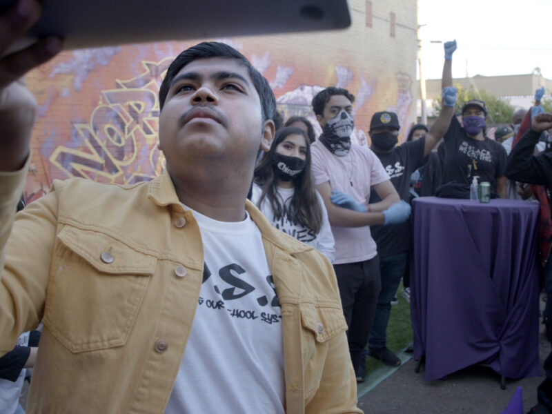 Still from "Homeroom" in which student Denilson Garibo holds up a laptop to point the webcam at a group of people behind him.