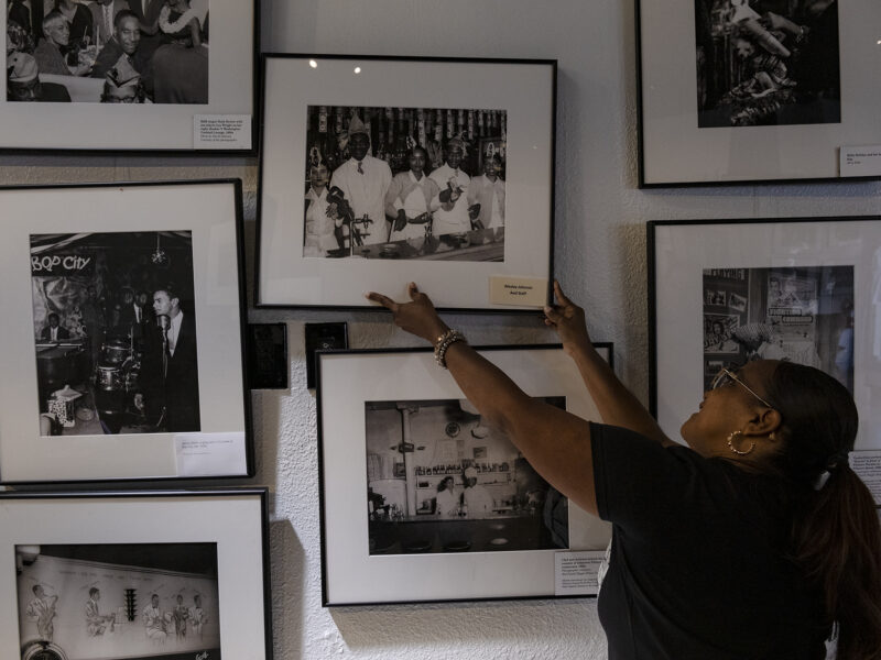 A woman with a long black ponytail reaches up to straighten the frame of one of many black and white photographs displayed in a closely spaced array on a wall in an art gallery.