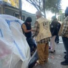 Three people wearing vests and jackets with city logos speak with a man standing in the entrance of a tent covered with plastic tarps on a city sidewalk.