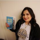 A woman stands against a wall holding a copy of Solito, Solita, a book about border-crossing youth.