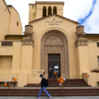 A person walks across the frame in front of a short set of stairs leading to the entrance of Laguna Honda Hospital. The hospital — a 780-bed facility on 62 acres in San Francisco’s Twin Peaks neighborhood — is facing challenges in fulfilling a Centers for Medicare and Medicaid Services mandate to move out all patients by Sept. 13, 2022, before it can apply to the centers for recertification.