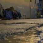 Two makeshift shelter structures are covered in a variety of mutely colored tarps to keep out the rain. They are built along a multi-colored wall that displays some graffiti. A parking sign nearby reads "Tow Away no parking any time" in all caps. In the foreground of the picture, puddles in the street are reflecting light from street lamps.