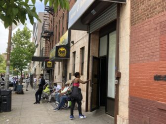 A woman wearing a red top opens the door to a brick building. Other people sit and stand near the entrance.