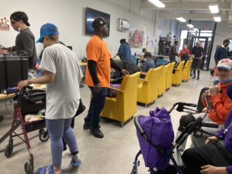 A line of yellow chairs with armrests features prominently in the center of a bustling waiting room.