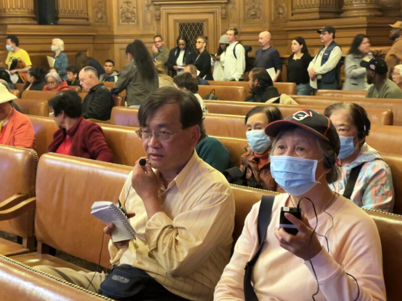 An interpreter speaks Cantonese into a device that transmits to listeners, with headsets, during a public meeting held in English.