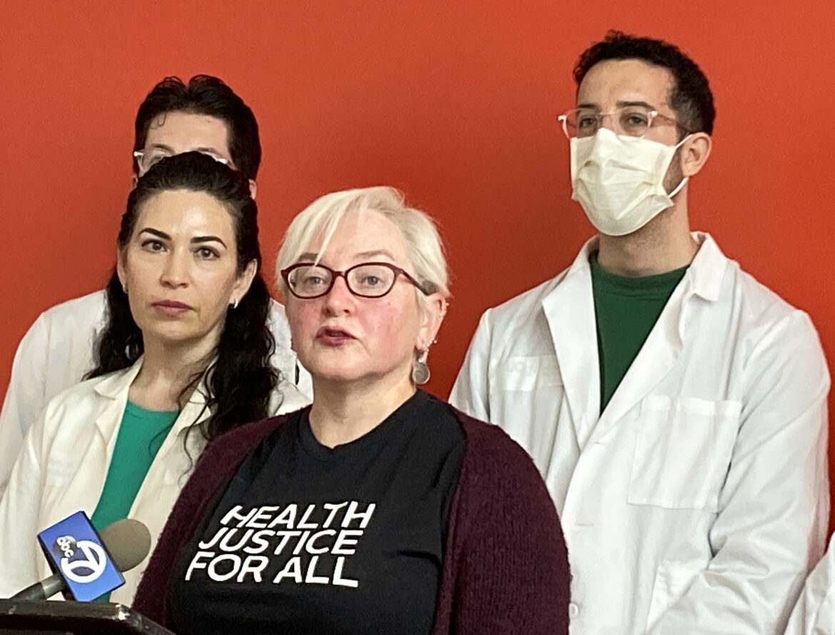 A woman with short, platinum blonde hair wearing glasses and a T-shirt printed with the words "Heath Justice for All" speaks into a microphone at a podium where she is standing near a woman and two men, all wearing white lab coats, in front of a red wall.