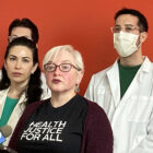 A woman with short, platinum blonde hair wearing glasses and a T-shirt printed with the words "Heath Justice for All" speaks into a microphone at a podium where she is standing near a woman and two men, all wearing white lab coats, in front of a red wall.