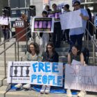 A group of people stand and sit holding signs on the steps of a court building. Some of the signs read: "open the courts, free our people," "speedy trial rights," "end the trial delay, free our people," "free our people," and "speedy trial violations." In the upper left background, woman standing at a lectern speaks into a microphone.