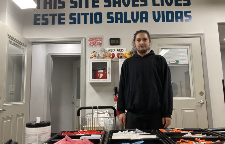 A man wearing dark clothing stands behind a table with syringes and trays of other medical supplies. The words "THIS SITE SAVES LIVES" above "ESTE SITIO SALVA VIDAS" are painted in blue in large letters high on the wall behind him.