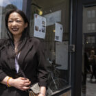 Susan Lefever, wearing a black jacket and crossbody purse, stands in front of the glass windows of a storefront. There are flyers posted on the glass behind her.