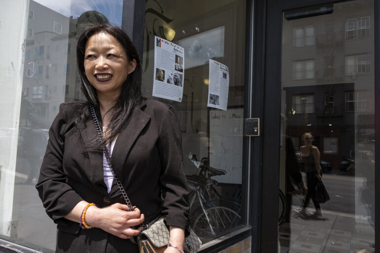 An Asian woman with long dark hair wearing a black blazer smiles standing in front of a glass storefront.