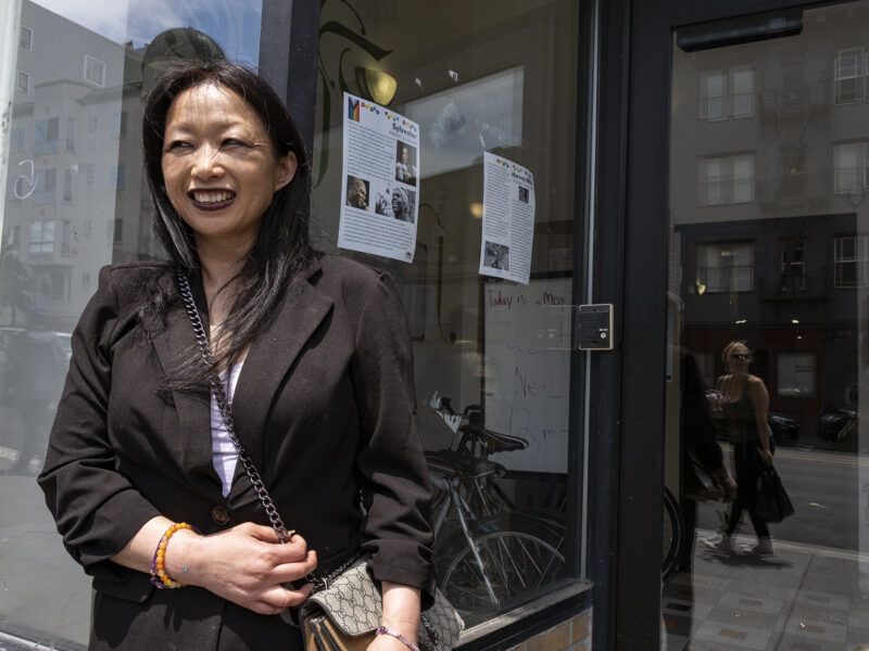 Susan Lefever, wearing a black jacket and crossbody purse, stands in front of the glass windows of a storefront. There are flyers posted on the glass behind her.