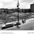 An aerial black and white photo displays an empty dirty lot surrounded by chain link fences. Two cars are parked next to the sidewalk in front of the lot. Behind the cars, a group of three people stand around a small table.