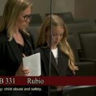 A girl dressed in black stands at a lectern where she is speaking. Her mother, also dressed in black, stands beside her. A few people sit behind them in theater-style seats.