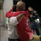 An adult forcibly picks up a boy wearing a red sweatshirt with a hood. Their faces are turned away. A police officer in uniform walks in the background.