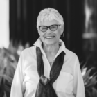 Black and white image of a woman with short silver hair wearing dark-framed eyeglasses, a light colored blouse and a dark scarf standing in front of leafy foliage. The background is slightly blurred.