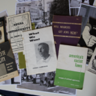 Eight historical booklets are spread out on top of several black and white photos of Black Americans in San Francisco protesting for civil rights in the 1960s. The pamphlet at the center of the table is titled "What We Want" and has a black-and-white photo of it's author Stokely Carmichael, who later changed his name to Kwame Ture. To its right is a lime green and white pamphlet called "America's Racist Laws" by Herbert Aptheker. Other pamphlet titles include "Behind the Lynching of Emmet Louis Till," ""Complete Equality: Democracy and the Negroes," and "Will the Negro Get Jobs Now?".