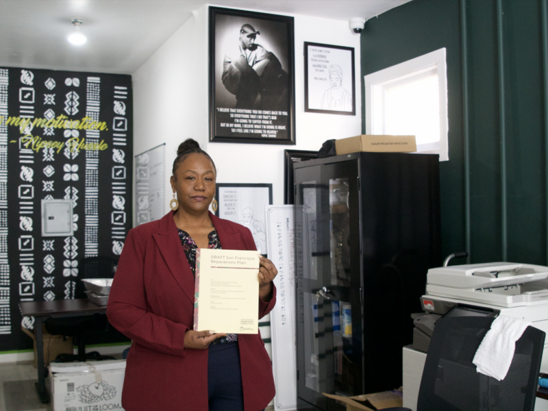 An African American woman in a burgundy jacket and blue pants is standing in an office space with several posters on the wall behind her. She holds a cream-colored booklet with the words "DRAFT San Francisco Reparations Plan" written across the top.