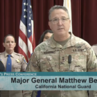 A man in fatigues speaks into a microphone at a news conference. He is surrounded by other people in police and law enforcement uniforms. They stand in front of flags and a burgundy curtain.