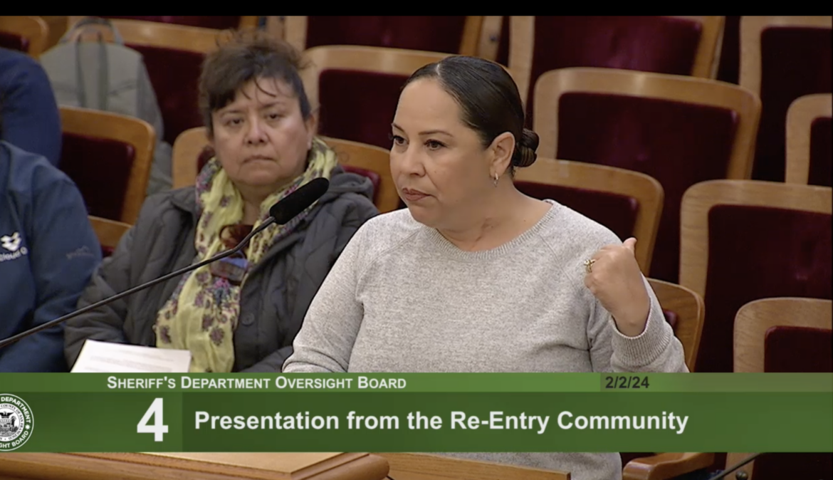 A woman wearing a long-sleeved, gray top and wearing her dark hair pulled back in a bun stands at a podium speaking into a microphone. Another woman appears in the frame seated behind her. This image is a screen-grab from a TV broadcast, and there is a green banner across the bottom of the frame with the following text: "Sheriff's Department Oversight Board" above "4: Presentation From the Re-Entry Community"