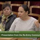 A woman wearing a long-sleeved, gray top and wearing her dark hair pulled back in a bun stands at a podium speaking into a microphone. Another woman appears in the frame seated behind her. This image is a screen-grab from a TV broadcast, and there is a green banner across the bottom of the frame with the following text: "Sheriff's Department Oversight Board" above "4: Presentation From the Re-Entry Community"