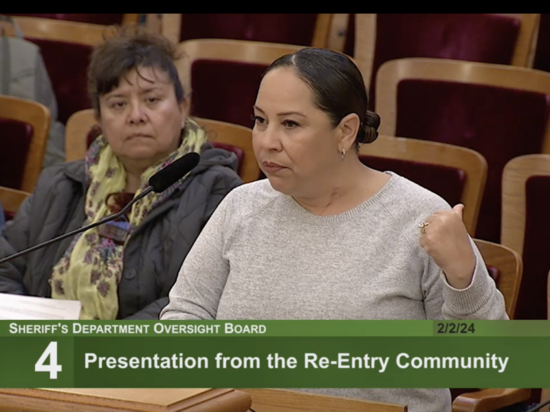 A woman wearing a long-sleeved, gray top and wearing her dark hair pulled back in a bun stands at a podium speaking into a microphone. Another woman appears in the frame seated behind her. This image is a screen-grab from a TV broadcast, and there is a green banner across the bottom of the frame with the following text: "Sheriff's Department Oversight Board" above "4: Presentation From the Re-Entry Community"